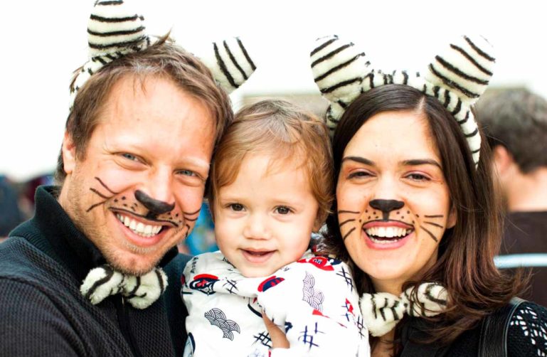 Parents with cat whisker face paint hold young child during Purim celebration