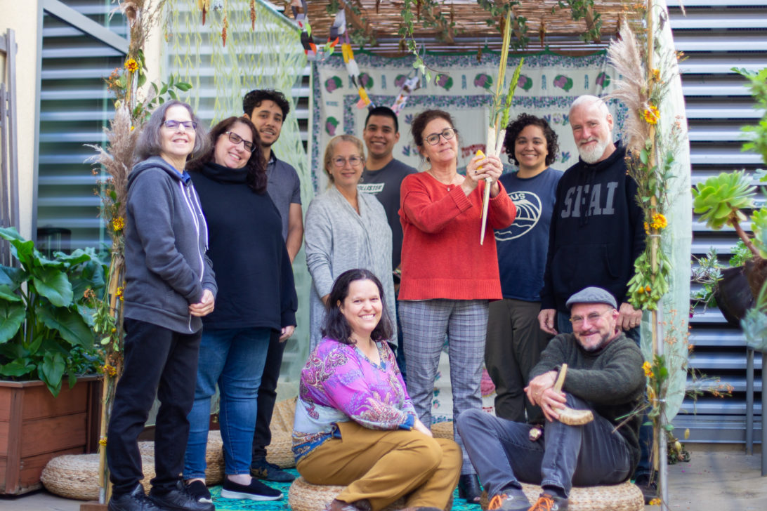 A group of staff gather around a sukkah while a woman in the center holds a lulav and etrog.